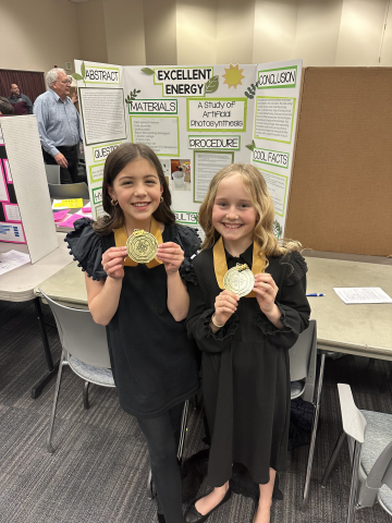 Riley and Ari showing their medals in front of their science fair project.